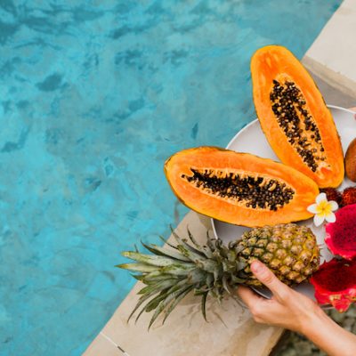 Woman holding plate of tasty tropical exotic fruits on the edge of pool, breakfast at luxury hotel. Slices of papaya, dragon fruit, passion fruit. Vegan and healthy food.