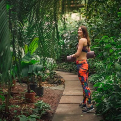 Full length portrait of beautiful smiling dreamy young woman in a colorful sporty outfit standing on a path, holding a mat at the botanical garden.Healthy lifestyle concept.